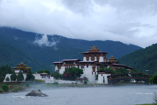 Traditional fortress-style building with white walls and ornate roofs beside a river, surrounded by misty green mountains