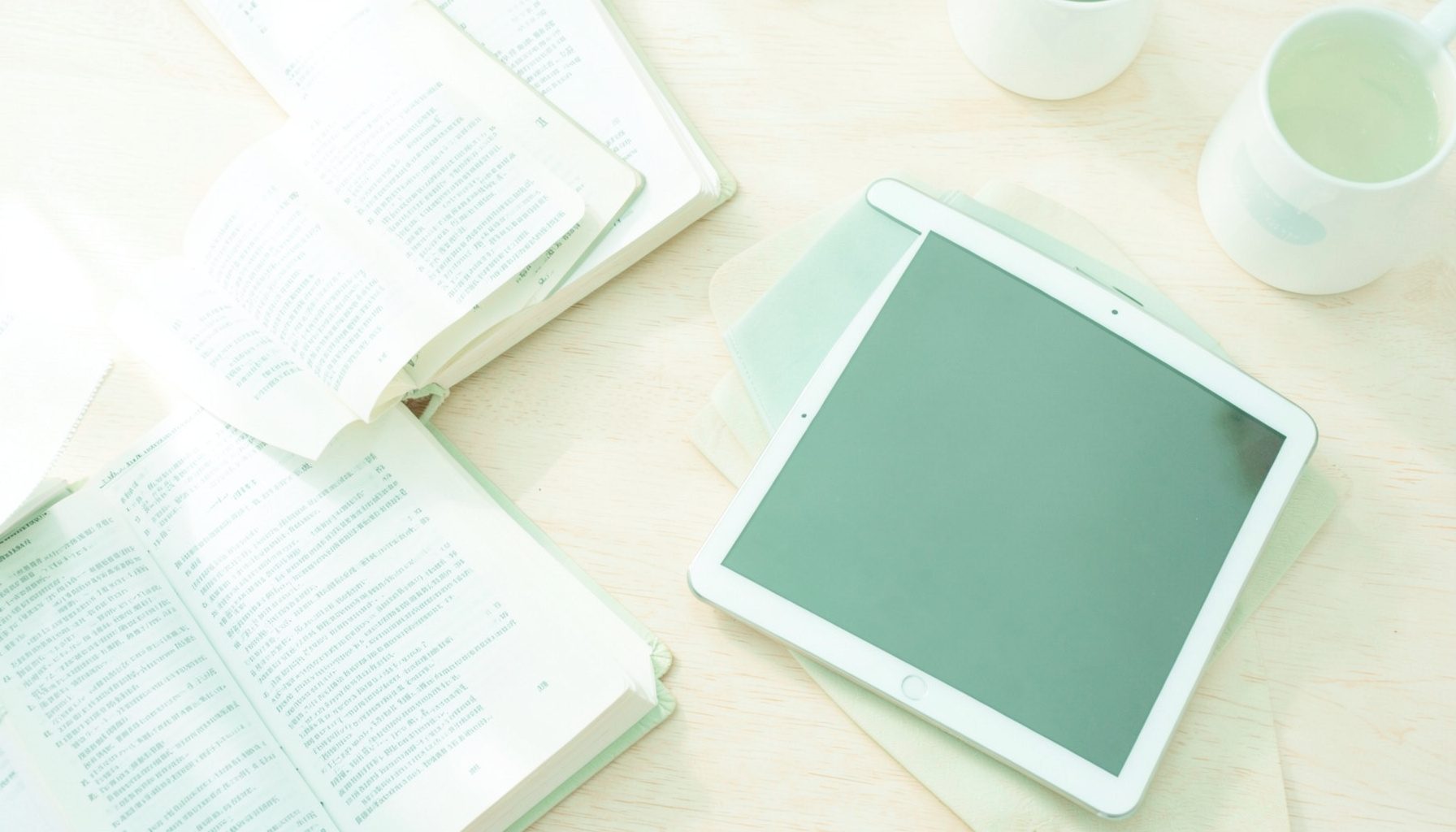 A bright, airy overhead view of open books and a tablet on a light wooden desk, with soft natural daylight and pale neutrals.